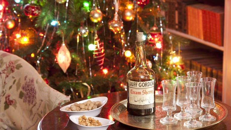 A bottle of cream sherry and five sherry schooner glasses set on a table next to an armchair with a decorated Christmas tree in the background